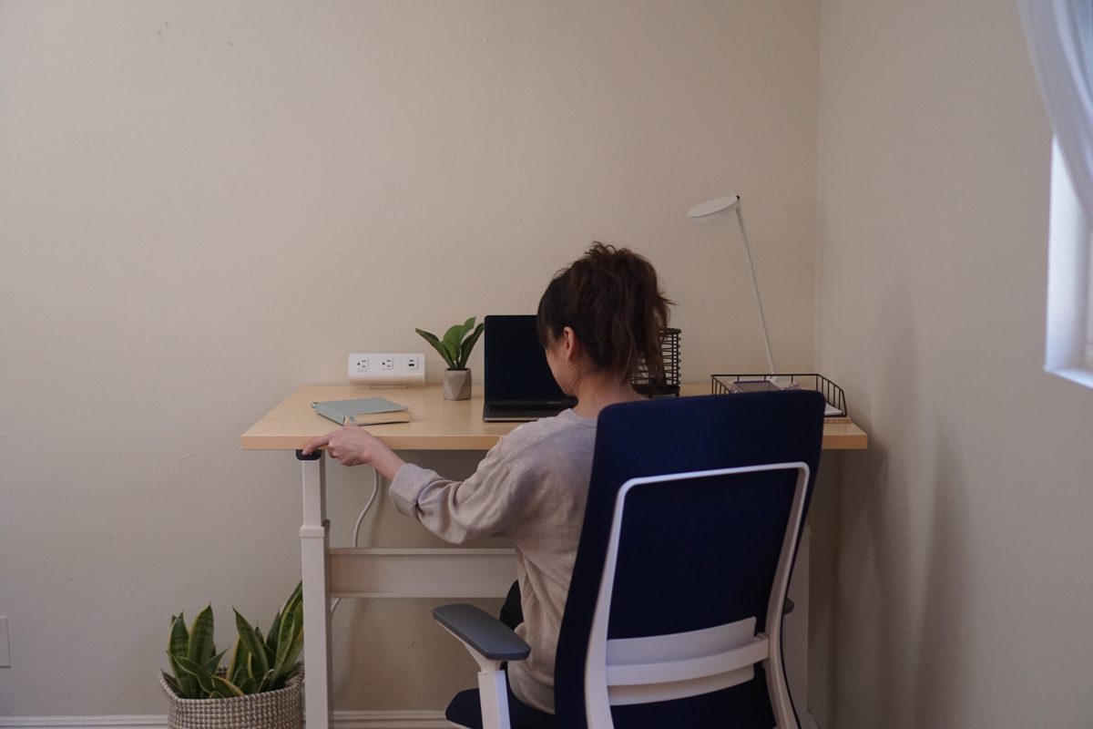 A woman seen from behind, working at a wooden desk in a corner, sitting in a navy Eurotech Elevate with a white frame