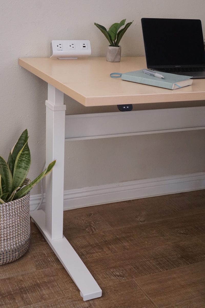 The end of a Bonita ET desk against a white wall, with its maple top, a white T-leg and foot on a dark brown floor, a laptop and a power strip on top, and a potted plant in a woven basket beside the leg