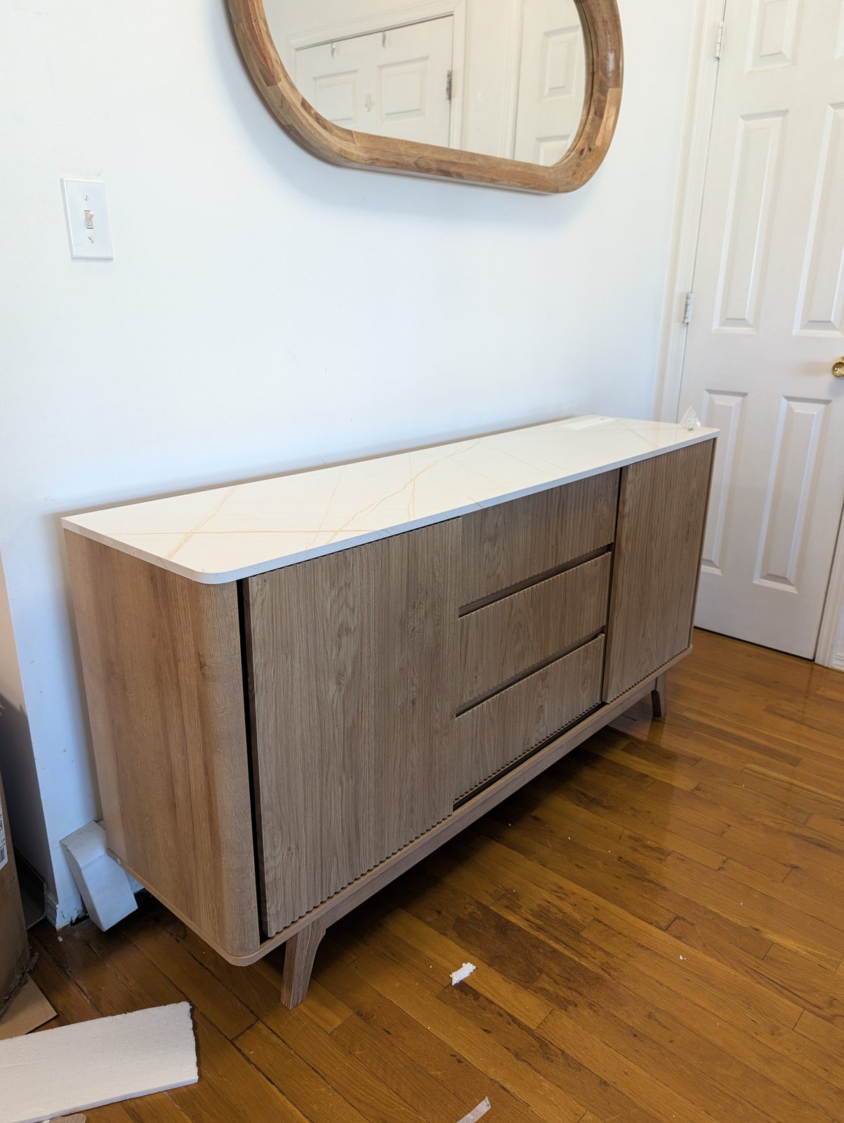 The finished light oak fluted sideboard with a white marble-look top under an oval wood-framed mirror, with bits of packing foam on the wood floor
