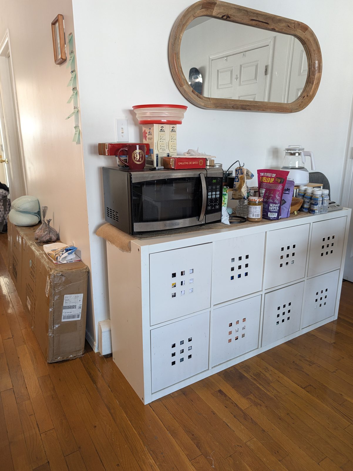 A white IKEA Kallax shelf with white cube inserts, a microwave and a pile of snacks and jars on top, under an oval wood-framed mirror, with a long cardboard box on the floor beside it
