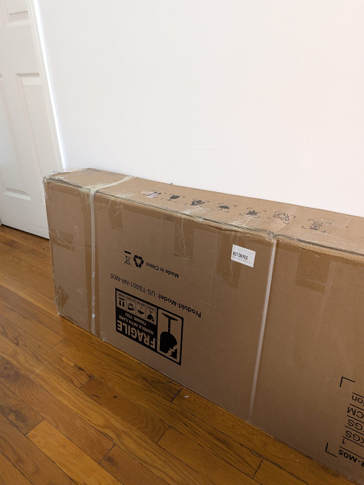 A long cardboard box leaning against a white wall on a wood floor, printed with a fragile label, beside a white door