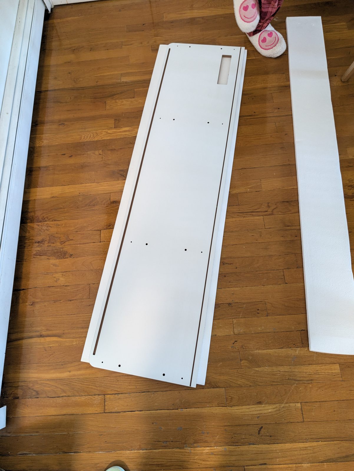 A long white pre-drilled panel lying on a wood floor beside a strip of white foam