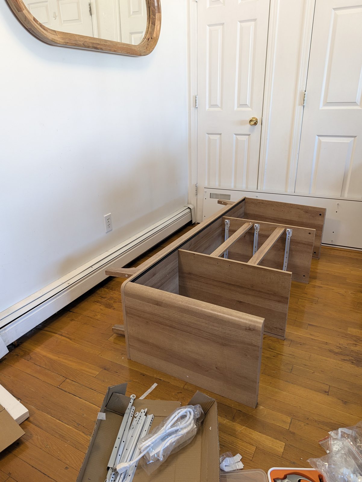 The partly built oak-look cabinet body lying on a wood floor with its rounded side panel, dividers and white metal drawer runners in place, a box of hardware and an orange tool case in front of it