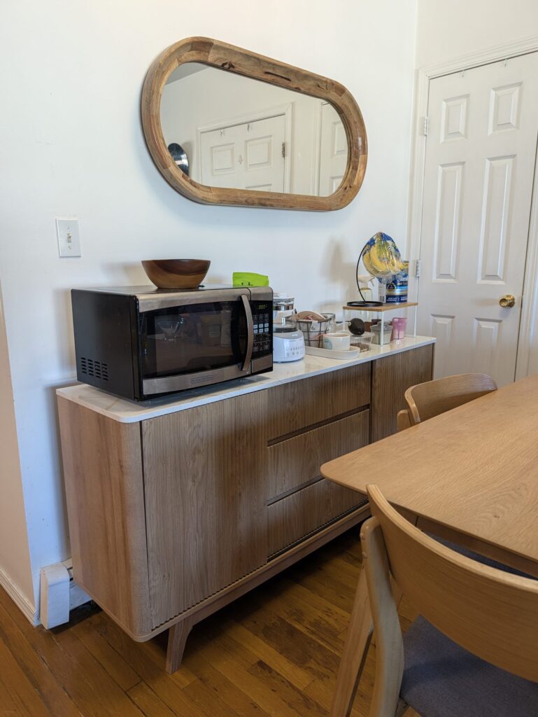 A light oak fluted sideboard with a white marble-look top under an oval wood-framed mirror, with a microwave, a kettle and kitchen things on top, beside a light wood dining table and chairs