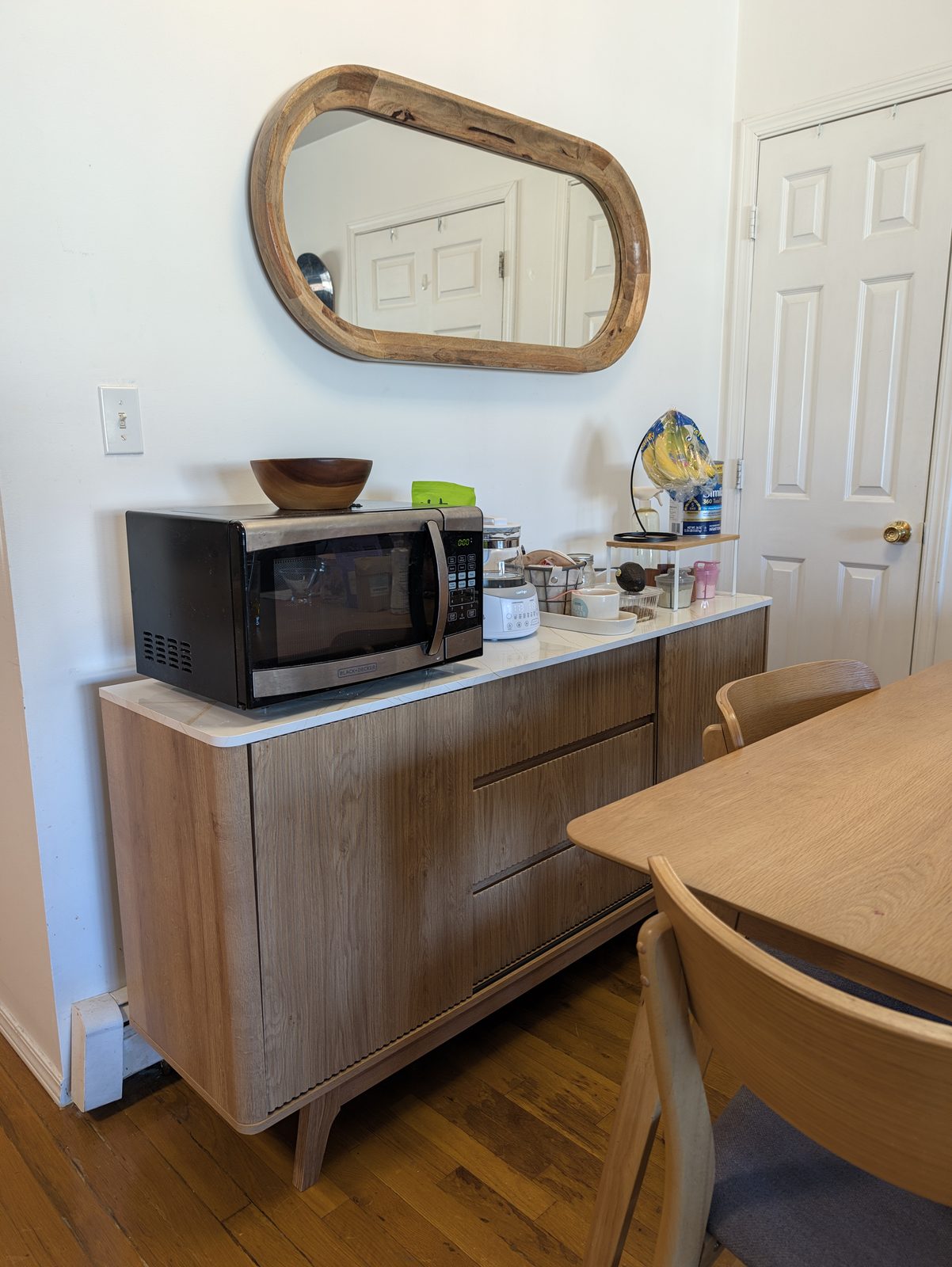 A light oak fluted sideboard with a white marble-look top under an oval wood-framed mirror, with a microwave, a kettle and kitchen things on top, beside a light wood dining table and chairs