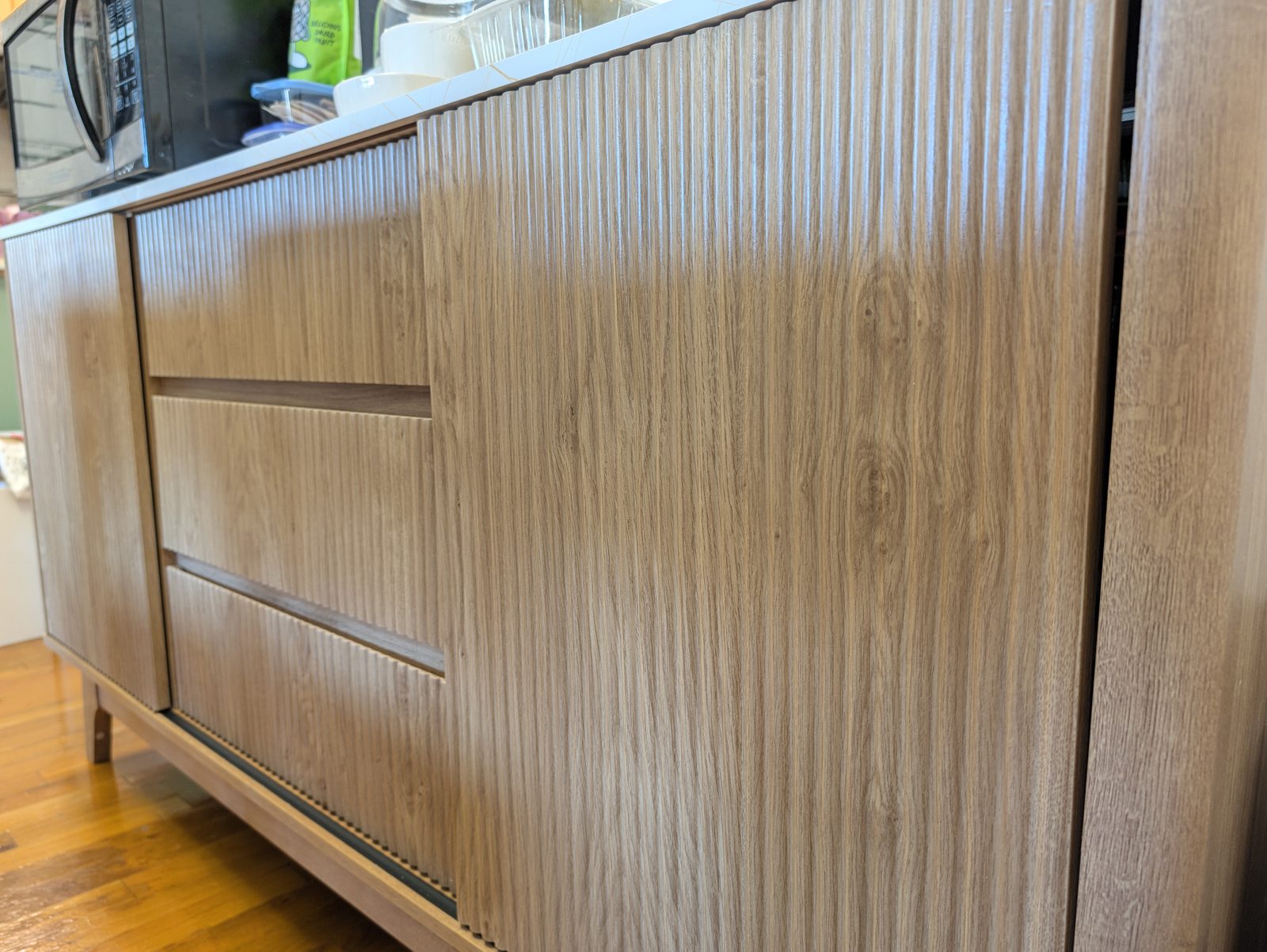 A fluted sliding door pushed open across the front of three fluted drawers on a light oak sideboard, with a microwave on the marble-look top