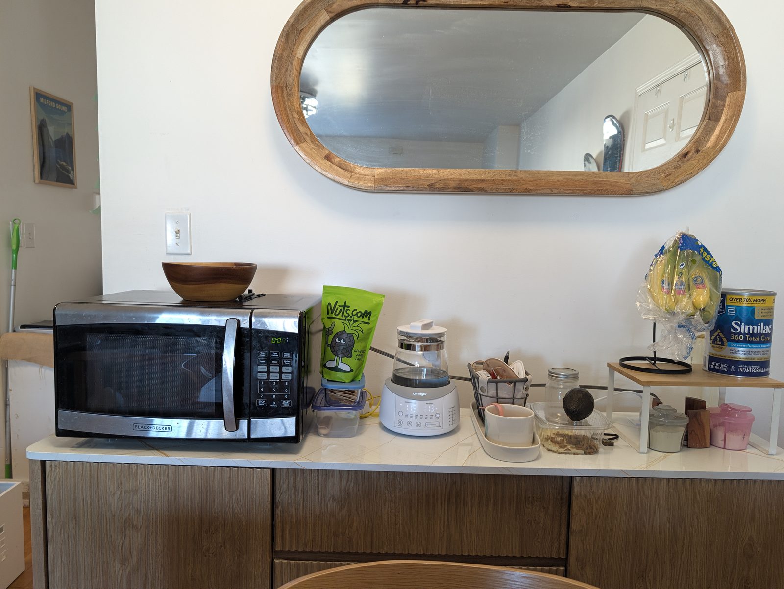 A marble-look sideboard top under an oval mirror holding a black and silver microwave with a wood bowl on it, a kettle, jars, a banana hook on a small riser and a can of baby formula