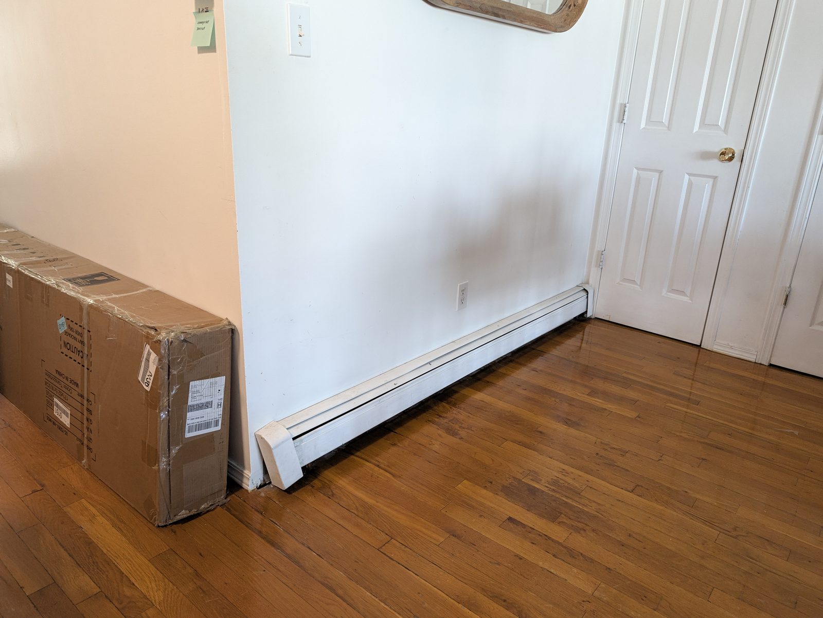 A bare white wall with a white baseboard heater along a wood floor, a wall outlet above it, a white door at the right and the long cardboard box on the left