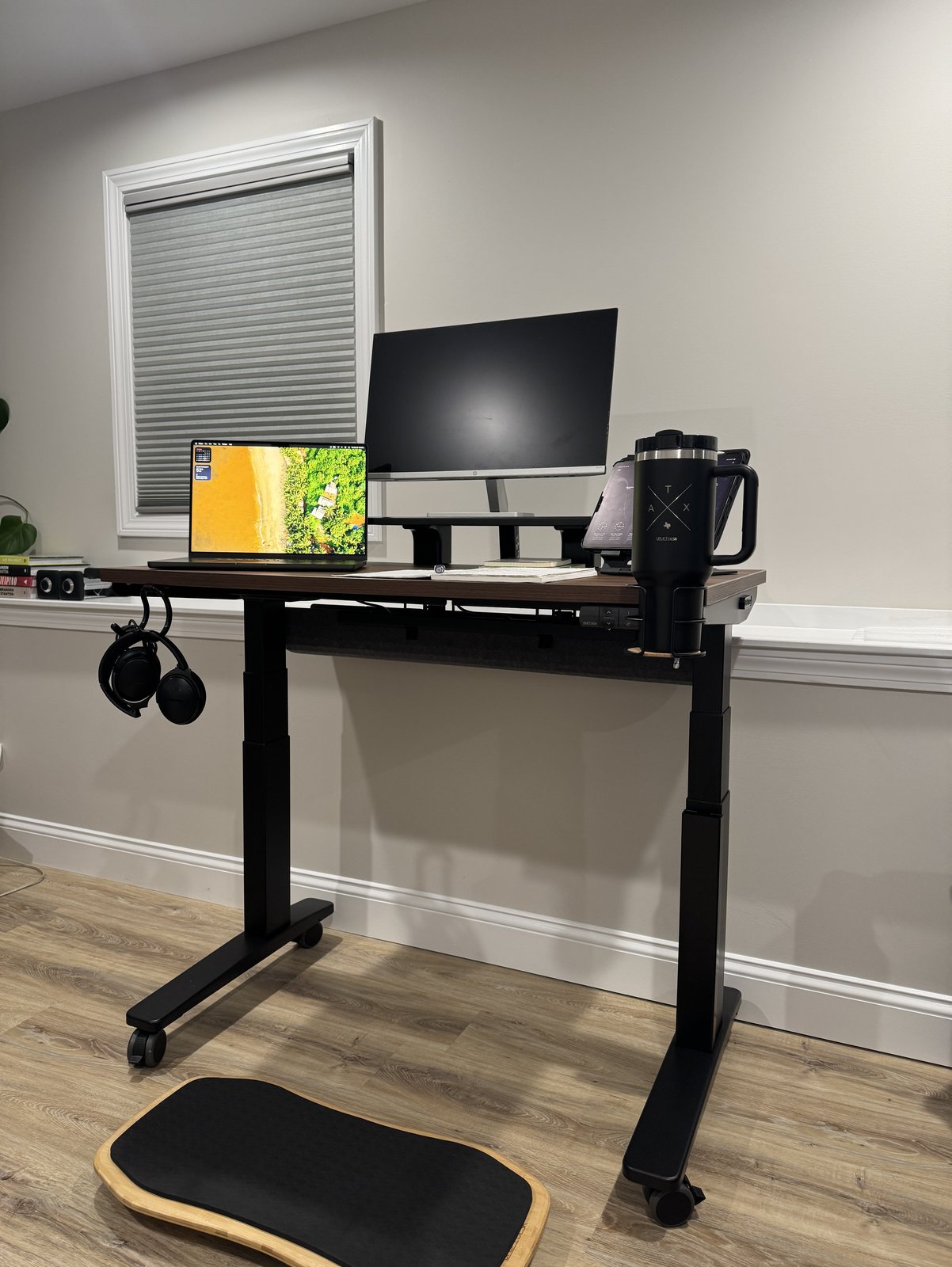 An Uplift V3 standing desk shown down to the floor, with a walnut top, black legs on casters, an HP monitor on a desk shelf, a laptop, an iPad on a tablet stand, a tumbler in a cup holder, headphones on a hook and a gray felt pouch under the back edge, and a bamboo Motion-X board on the floor in front of it