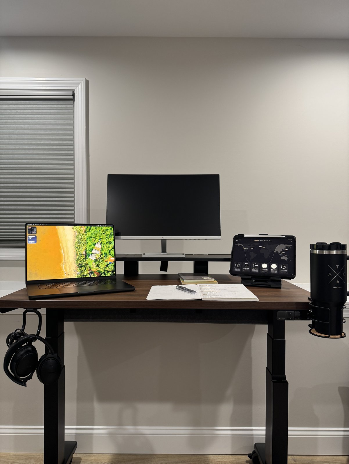 The whole front of an Uplift V3 desk at sitting height, with a walnut top on a black frame, a laptop, an HP monitor on a black desk shelf, an iPad on a tablet stand, an open notebook, headphones on a hook at the left end, the keypad under the front edge and a tumbler in a cup holder at the right end