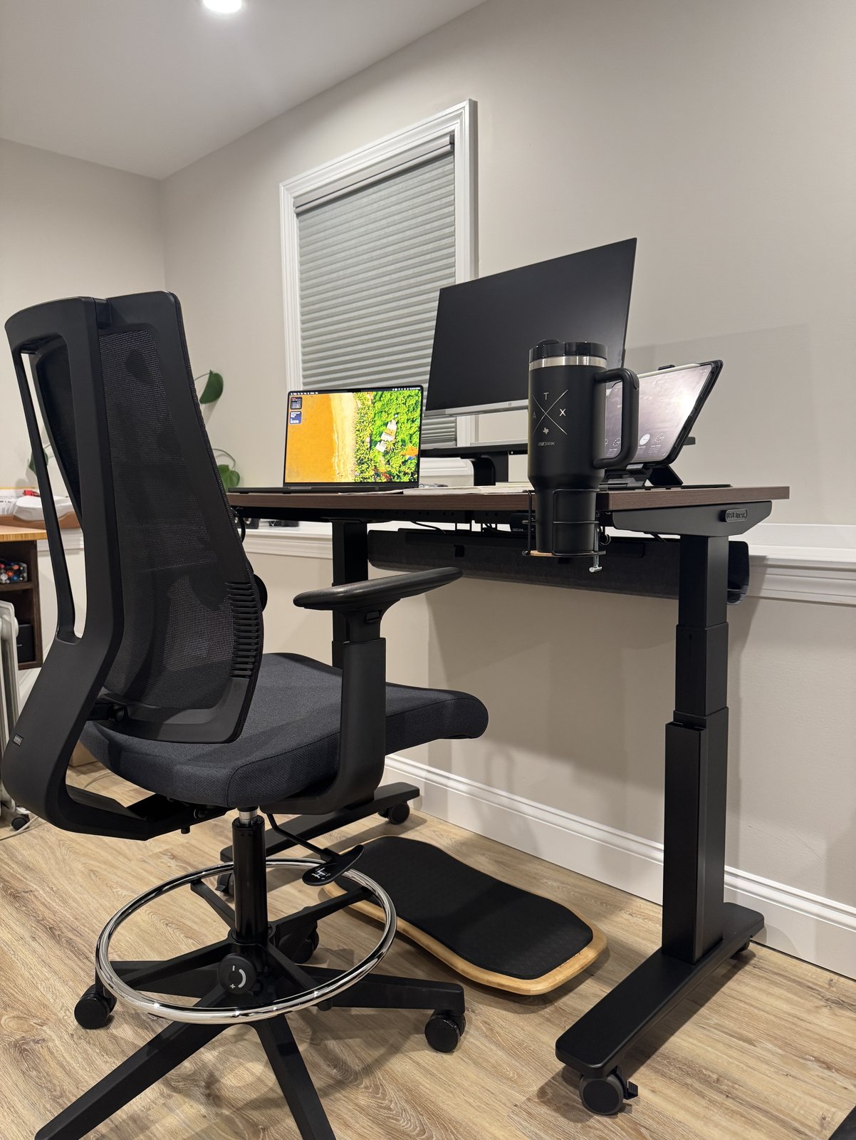 An Uplift Clarksville drafting chair with a mesh back and a chrome foot ring beside an Uplift V3 desk with a walnut top, with a bamboo Motion-X board on the floor under the front of the desk