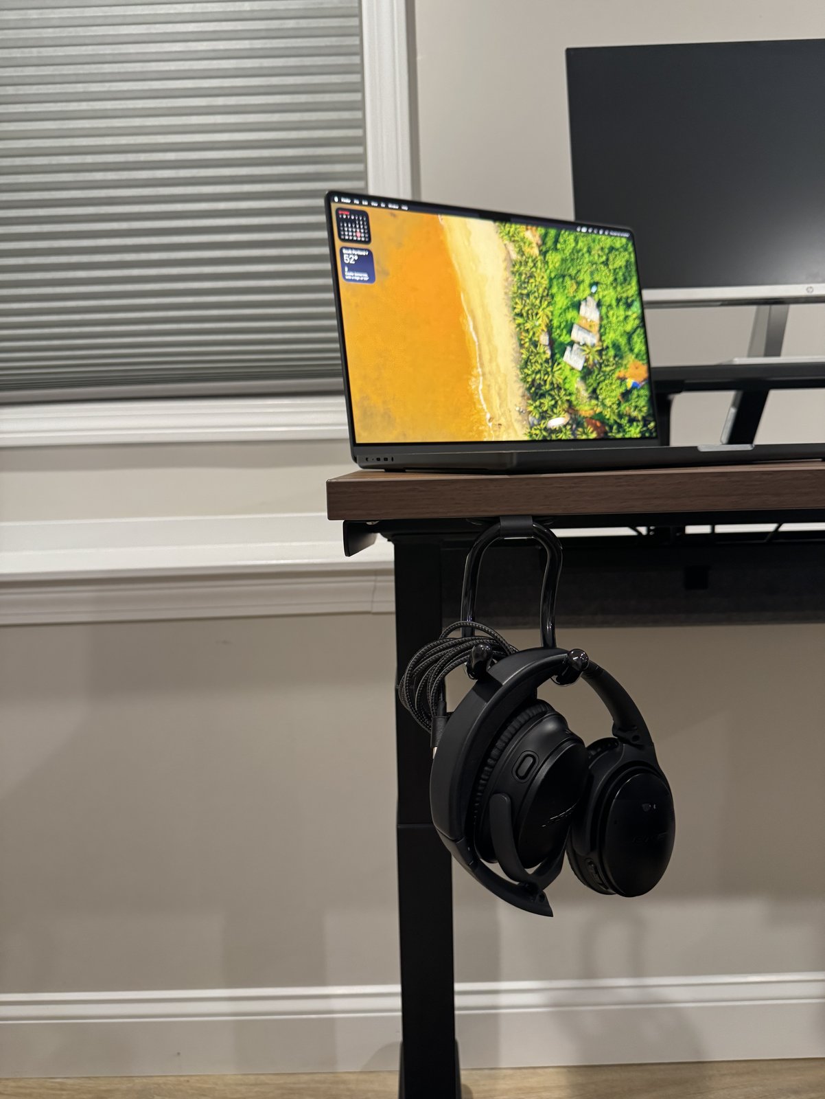 Headphones hanging from a black flexible hook clipped under the left edge of a walnut desk