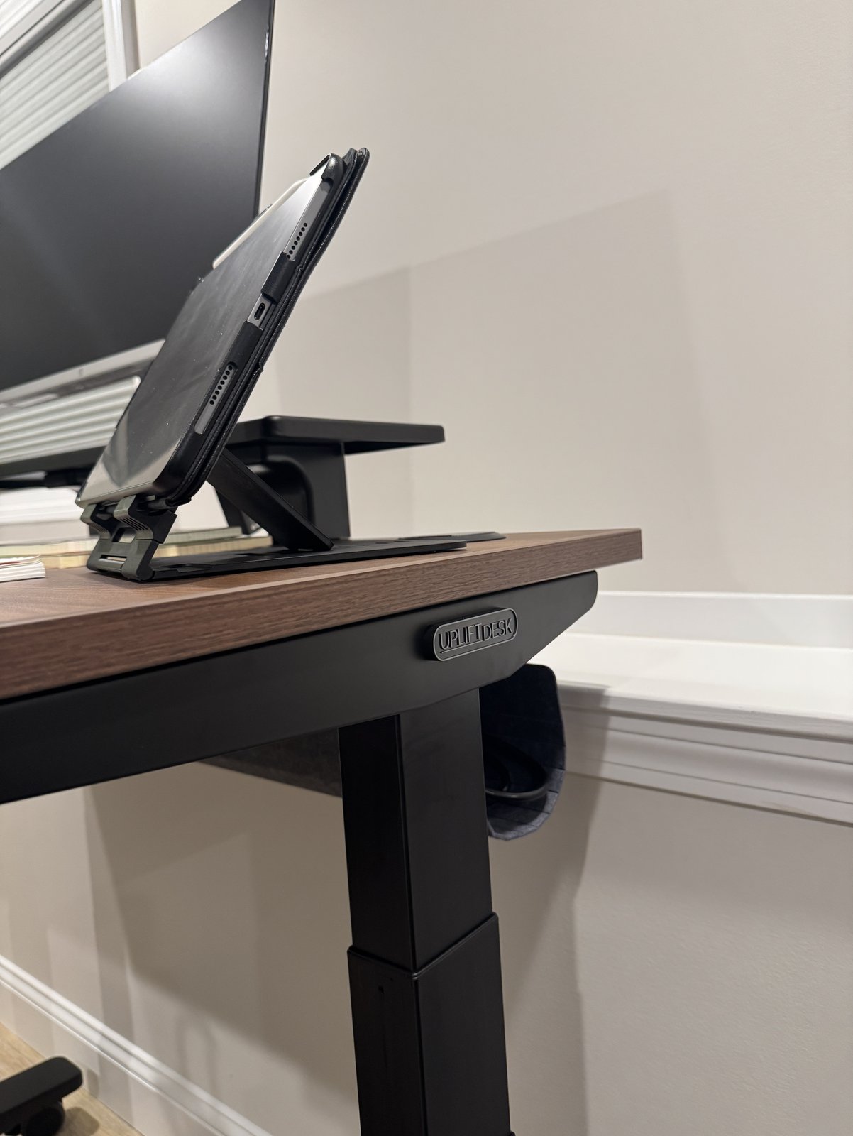 The right end of an Uplift V3 desk, showing the walnut laminate edge, an iPad on a tablet stand, the Uplift badge on the black frame and a gray felt pouch under the back