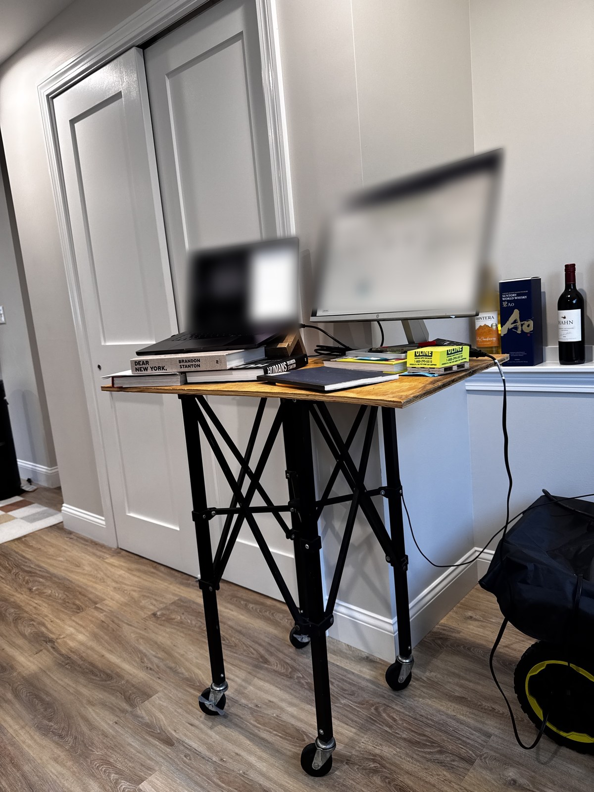 A homemade standing desk next to a closet door, made of a plywood top on a black folding stand with casters, with a laptop raised on a stack of books and a monitor beside it, both screens blurred