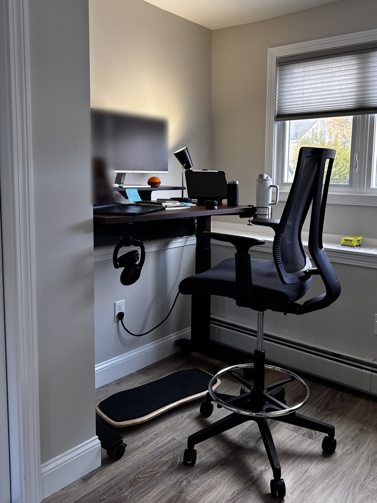An Uplift V3 standing desk with a walnut top and black frame on casters in a corner by a window, with a monitor on a desk shelf, a lamp, a tablet stand, a tumbler in a cup holder and headphones on a hook, a black drafting chair beside it and a bamboo Motion-X board on the floor