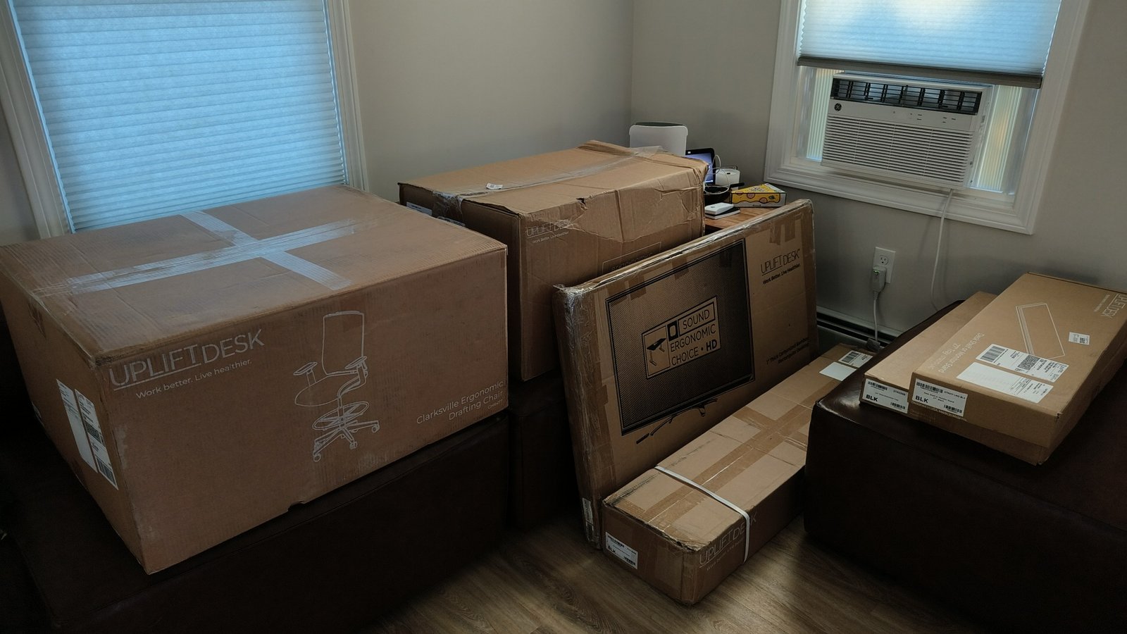 The first shipment's Uplift boxes, including the Clarksville drafting chair's box, the desktop's box, the frame's long box and two flat boxes for the desk shelf and extension tray, in the corner of a room with two windows, with no people in the photo