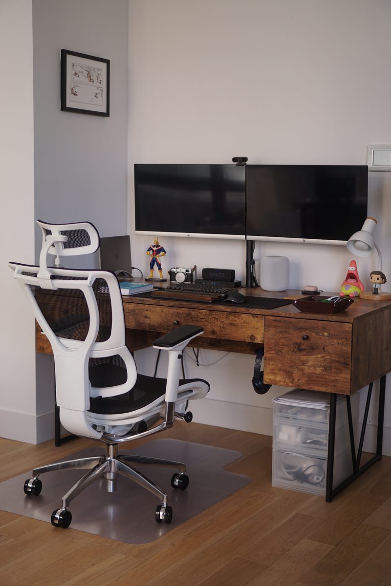 A white-framed Eurotech iOO with a mesh headrest, seen from the side and behind, at a wooden desk with drawers beneath two monitors