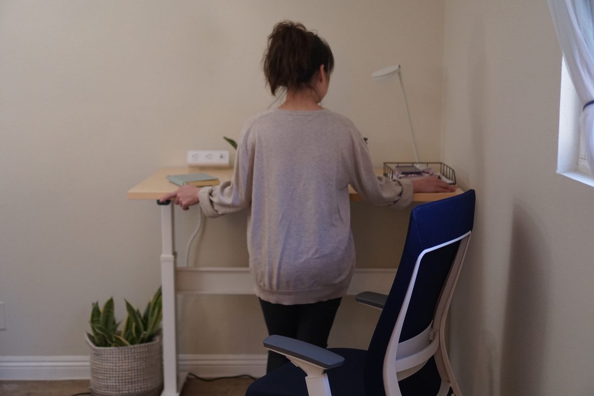 A woman seen from behind, standing at a raised RightAngle Bonita standing desk in the corner of a den with both hands on its maple top, a navy Eurotech Elevate pushed to one side and a potted plant in a woven basket by the desk leg